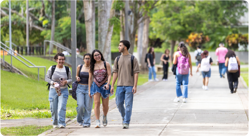 Estudiantes caminando juntos por un sendero arbolado del campus, conversando y sonriendo entre clases en la Universidad de Puerto Rico.