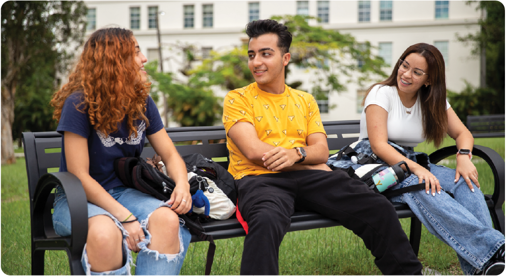 Tres estudiantes universitarios sentados en un banco al aire libre dentro del campus, conversando y compartiendo frente a un edificio universitario de UPR Cayey. Tres estudiantes universitarios sentados en un banco al aire libre dentro del campus, conversando y compartiendo frente a un edificio universitario de UPR Cayey.